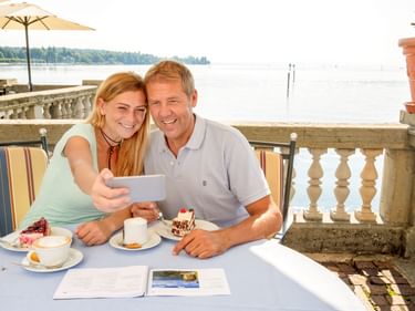 Lächelndes Paar auf Seeterrasse in Konstanz mit Reisebroschüre und Desserts beim Selfie. Bodensee und Ufer im Hintergrund sichtbar.