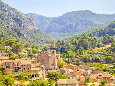 Dorf Valldemossa mit Steinkirche und traditionellen Häusern in grünem Tal, umgeben von bewaldeten Bergen unter klarem Himmel.