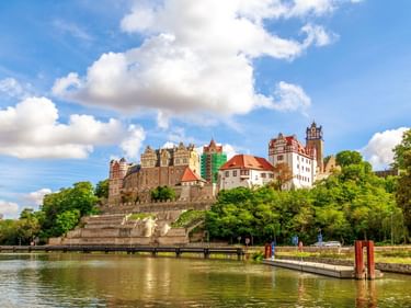 Schloss Bernburg mit historischen Gebäuden und Türmen auf einem Hügel über der Saale, umgeben von grünen Bäumen unter blauem Himmel mit Wolken.