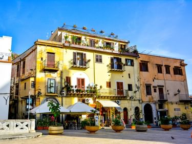Bunte Gebäude um einen Platz in Monreale mit Straßencafés. Gelbe und beige Fassaden mit Balkonen unter blauem Himmel.