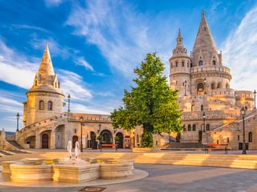 Fischerbastei in Budapest mit weißen Steintürmen und Bögen. Eine Person in Weiß überquert den Platz mit einem Brunnen im Vordergrund.