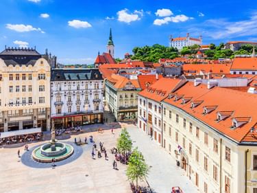 Bratislava Altstadtplatz mit Brunnen, historischen Gebäuden mit orangen Dächern und Burg auf dem Hügel unter blauem Himmel.