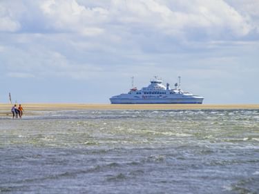 Weiß-blaue Fähre vor Sylt mit Menschen im Watt im Vordergrund. Sandstrand und bewölkter Himmel im Hintergrund sichtbar.