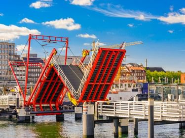 Rote Hörnbrücke Klappbrücke in Kiel hochgeklappt mit Booten im Hafen. Weiße Fußgängerstege und Stadtgebäude unter blauem Himmel sichtbar.