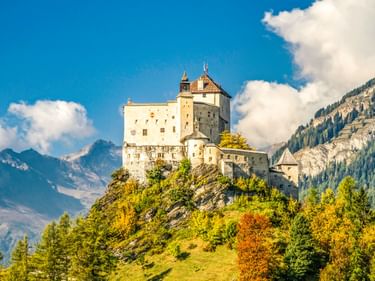 Schloss Tarasp thront auf einem Hügel, umgeben von Herbstlaub, mit Bergen und blauem Himmel im Hintergrund.