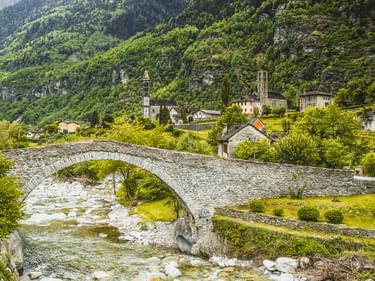 Alte Steinbogenbrücke über einen Gebirgsfluss in Giornico, Tessin. Dorf mit Kirchtürmen eingebettet vor bewaldeten Berghängen.