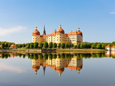 Schloss Moritzburg mit orangen Kuppeln und gelber Fassade spiegelt sich im ruhigen Seewasser. Bäume säumen das Ufer unter blauem Himmel.