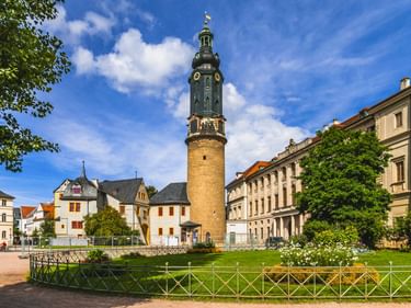 Historischer Turm mit grüner Kuppel am Stadtschloss Weimar ragt über umliegende Gebäude. Grüner Park mit Ziergitter im Vordergrund unter blauem Himmel.