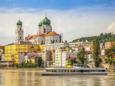 Der Passauer Dom St. Stephan mit grünen Zwiebeltürmen überragt bunte Gebäude entlang der Donau. Ein Passagierschiff fährt auf dem Wasser.