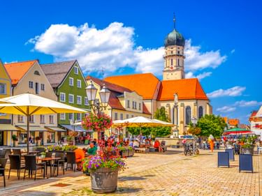 Stadtplatz in Schongau mit der Mariae Himmelfahrt Kirche mit Zwiebelturm, bunten historischen Gebäuden, Straßencafés und Blumenkübeln.
