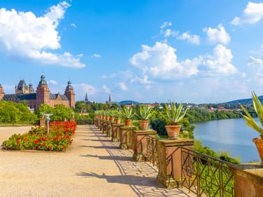 Terrace with potted plants and red flowers overlooking Schloss Johannisburg and the Main River in Aschaffenburg under a blue sky.
