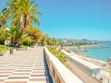 Gepflasterte Promenade mit geometrischem Muster und Palmen entlang der Mittelmeerküste in Sanremo. Strand und Berge im Hintergrund.