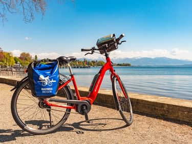 Rotes E-Bike mit blauer Radweg Reisen Fahrradtasche am Bodenseeufer in Kressbronn geparkt, mit Bergen im Hintergrund.