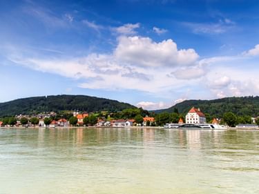 View of Obernzell on the Danube River with a white baroque building, red-roofed houses, and forested hills under a blue sky with clouds.