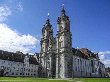 Stiftskirche St. Gallen mit barocken Zwillingstürmen und verzierter Fassade, umgeben von historischen Gebäuden und grüner Wiese.
