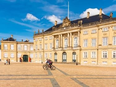 Radfahrer überquert den Kopfsteinpflasterplatz am Schloss Amalienborg in Kopenhagen. Die gelbe Rokoko-Palastfassade mit Säulen unter blauem Himmel.