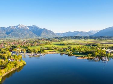 Luftaufnahme des Hafens Feldwies am Chiemsee mit Segelbooten, grünen Wiesen und den Chiemgauer Alpen im Hintergrund unter blauem Himmel.