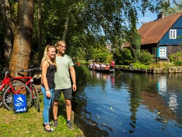 Zwei Radfahrer stehen mit Fahrrädern an der Spree unter großen Bäumen. Ein blaues traditionelles Haus und ein Boot mit Passagieren sind im Hintergrund sichtbar.