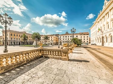 Piazza d'Italia in Sassari mit verzierter Balustrade, historischen Gebäuden, Palmen und verzierten Straßenlaternen unter blauem Himmel mit weißen Wolken.