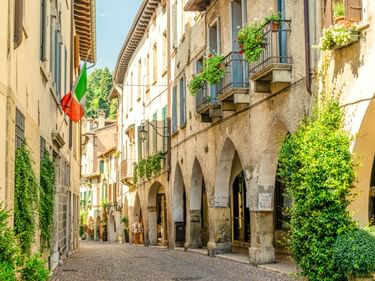 Schmale Kopfsteinpflastergasse in Asolo mit Steinbögen, verwitterten Gebäuden mit Balkonen, grünen Pflanzen und italienischer Flagge an einem Fenster.