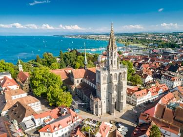 Luftaufnahme des Konstanzer Münsters mit hohem gotischen Turm über rotgedeckten Gebäuden. Bodensee mit Segelbooten und fernem Ufer sichtbar.