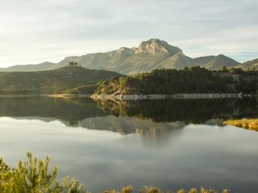Ruhiger See von Beniarres spiegelt Berge und bewaldete Uferlinie. Ein markanter Felsgipfel erhebt sich im Hintergrund im weichen Abendlicht.