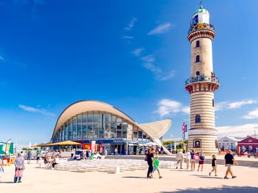 Das markante weiße Teepott-Gebäude mit geschwungenem Dach und hoher gestreifter Leuchtturm im Rostocker Hafen, mit Besuchern auf dem Platz.