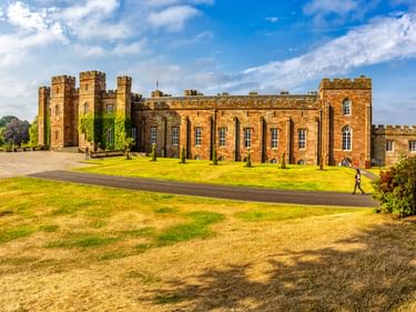 Scone Palace in Perth, ein Schloss aus rotem Sandstein mit Zinnen und hohen Fenstern, umgeben von gepflegten Rasenflächen und Zypressen unter blauem Himmel.