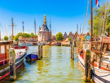 Hafen in Hoorn mit traditionellen Booten an Holzpfählen. Historischer Turm und Giebelhäuser unter blauem Himmel sichtbar.