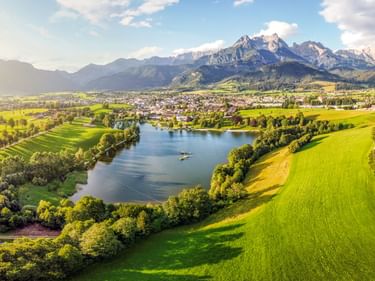 Luftaufnahme von Saalfelden mit grünen Wiesen, einem See und schneebedeckten Bergen im Hintergrund unter blauem Himmel mit Wolken.