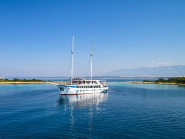 Weiße Motoryacht MS Aria mit zwei Masten vor Anker in ruhigem blauen Wasser nahe der grünen kroatischen Küste unter klarem Himmel.