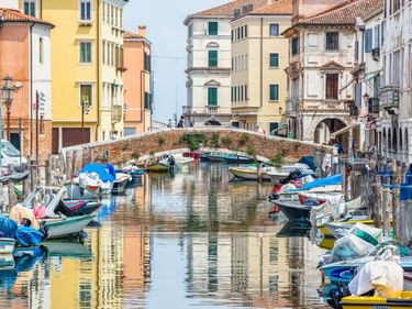 Kanal in Chioggia mit bunten Booten an Holzpfählen. Eine Backsteinbrücke überspannt das Wasser, pastellfarbene Gebäude säumen beide Seiten.