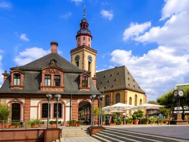 Katharinenkirche mit gelbem Turm und historischem rotbraunem Gebäude auf Frankfurter Platz. Weiße Sonnenschirme und Pflanzkübel im Vordergrund.
