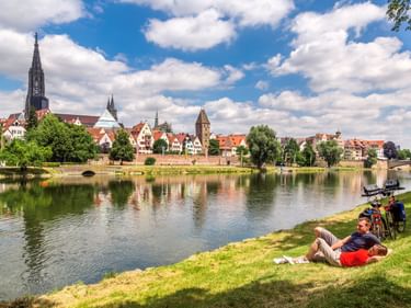 Radfahrer ruht auf Wiese an der Donau mit Tourenrad. Ulms historische Skyline mit Münsterturm spiegelt sich im ruhigen Wasser.