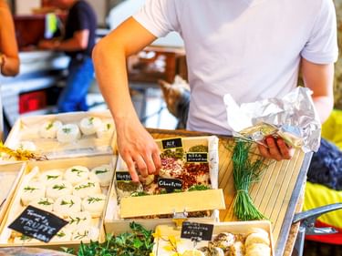 Verkäufer im weißen Hemd arrangiert Käseprodukte an einem Marktstand in Orange. Auslage mit verschiedenen Käsesorten und frischen Kräutern.