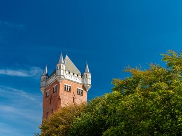 Esbjerg Wasserturm mit rotem Ziegelsockel und weißer Zinnenspitze mit vier Türmchen, umgeben von grünen Bäumen vor blauem Himmel.