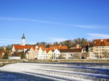 Flussufer von Landsberg am Lech mit bunten historischen Gebäuden, roten Ziegeldächern und Kirchturm. Ein Stufenwehr durchquert den Lech.
