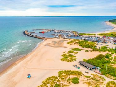 Luftaufnahme des Hafens von Hornbæk mit Marina und Sandstrand. Weiße Boote am Wellenbrecher, grüne Vegetation und blaues Meer sichtbar.