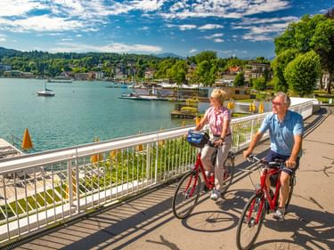 Zwei Radfahrer auf roten Rädern fahren entlang eines Seewegs am Wörthersee. Der Weg hat weiße Geländer, Boote und Gebäude sind über dem Wasser sichtbar.