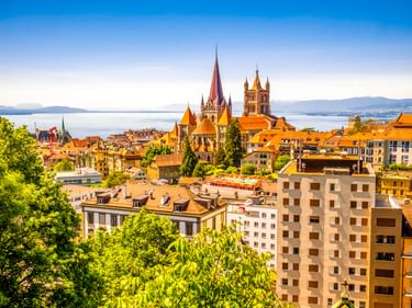 Luftaufnahme von Lausanne mit den Türmen der Kathedrale Notre-Dame über orangefarbenen Dächern. Genfersee und Berge im Hintergrund.