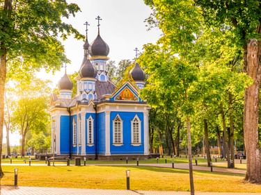Blue and white Orthodox church with dark onion domes and crosses in Druskininkai, surrounded by green trees in a park setting.