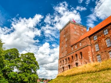 Schloss Koldinghus in Kolding mit rotem Backsteinturm und orangefarbenem Ziegeldach auf einem Grashügel unter blauem Himmel mit weißen Wolken.