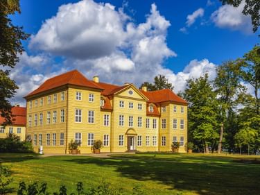 Schloss Mirow, ein dreistöckiges gelbes Barockschloss mit rotem Ziegeldach, umgeben von grünem Rasen und Bäumen unter blauem Himmel.