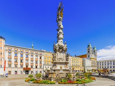 Linz Hauptplatz with ornate Trinity Column in center, surrounded by colorful historic buildings and a church with twin towers under blue sky.