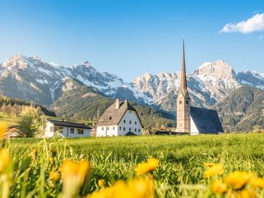 Kirche mit hohem Turm und traditionelle Häuser in Maria Alm, Österreich, eingerahmt von gelben Wildblumen und schneebedeckten Alpengipfeln.