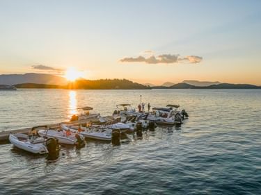 Mehrere weiße Motorboote am Steg bei Sonnenuntergang in Griechenland. Die Sonne geht hinter Inseln unter und wirft goldenes Licht.