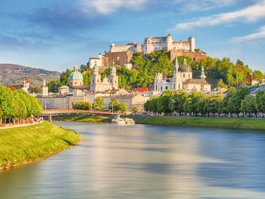 Festung Hohensalzburg auf einem Hügel über Salzburgs Altstadt mit barocken Kirchen und Kuppeln. Die Salzach fließt im Vordergrund.
