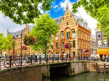 Brücke über Amsterdamer Gracht mit Fahrrädern, verzierten orangefarbenen Backsteingebäuden, hängenden Blumenkörben und grünen Bäumen unter blauem Himmel.