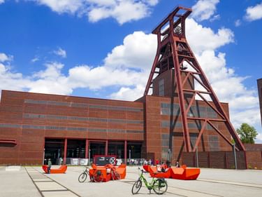 Zeche Zollverein Industriekomplex mit ikonischem roten Förderturm und Backsteingebäuden. Orange Sitzmöbel und Fahrräder auf dem Platz.