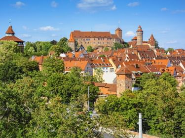 Blick auf Nürnbergs Altstadt mit roten Ziegeldächern, mittelalterlicher Stadtmauer, Türmen und Burg auf dem Hügel, umgeben von grünen Bäumen.